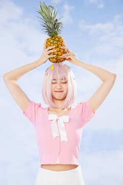 Playful young woman with pink hair holding pineapple on head against blue sky