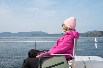 Fototapeta premium Woman in vivid pink jacket rests on catamaran while watching Pacific Ocean and rugged Kamchatka coast. Sea adventure, relaxation, nature tourism, travel lifestyle.