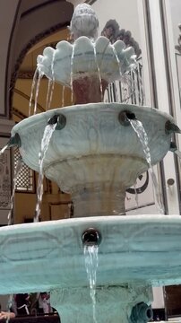 Close-up view of a traditional ornate marble fountain with multiple tiers and cascading streams of water. Decorative Islamic architectural elements and Arabic calligraphy are visible in the background