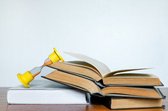Open books stacked on a desk with a fallen hourglass resting on the pages. Time is running out, symbolizing exam pressure, deadlines, and university studies.