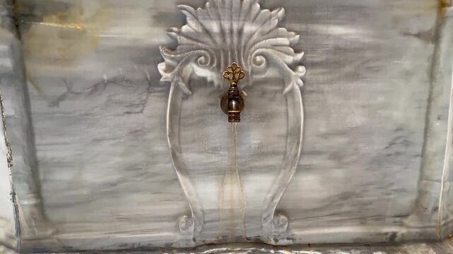 Close-up view of an ornate traditional brass faucet mounted on a decorative white marble fountain. The fountain features intricate carved floral and shell motifs, reflecting traditional Ottoman and Is