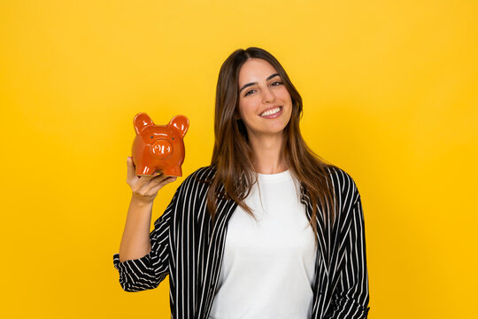 Young woman smiling and presenting a piggy bank, symbolizing savings, finance, investment, and future planning, standing against a bright yellow studio background with copy space