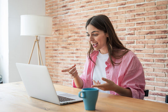 Young woman working from a bright home office, smiling and engaging in a lively video call on her laptop. Casual, positive remote work scene with natural light, coffee mug and exposed brick wall.