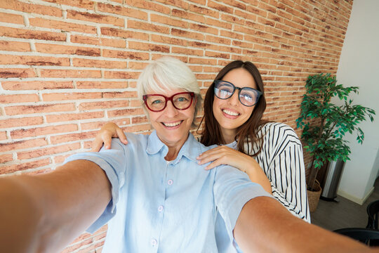 Happy grandmother and granddaughter taking a selfie together at home against a brick wall