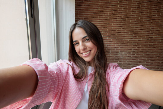 A smiling young woman takes a relaxed selfie by a sunlit window in a pink striped shirt, conveying warmth, confidence and casual lifestyle with a modern, cozy indoor and well‑being vibe.