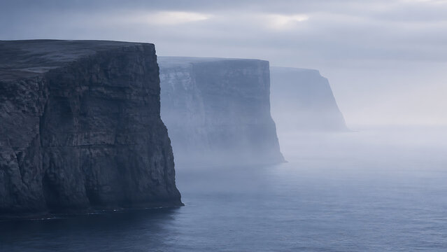 Tall dark rocky sea cliffs rising from the ocean under a gloomy overcast sky with thick mist clinging to the base of the stone formations