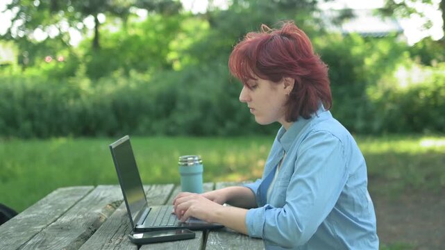 Ginger female typing on laptop keyboard in summer park.