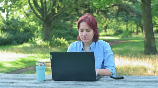 A young redhead woman sitting at a wooden table in a green park and opening her laptop. Concept of remote work, digital nomad lifestyle, outdoor office, and freelance freedom during summer.