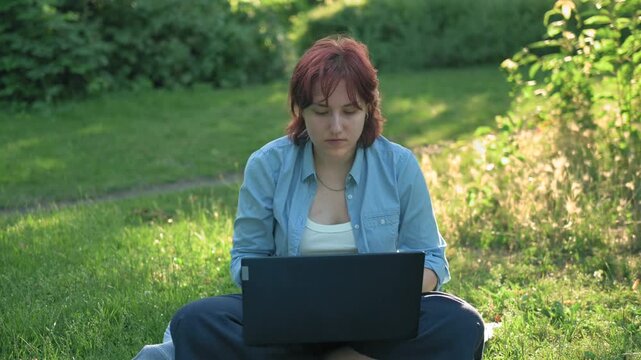 Focused ginger student preparing for exams on laptop on park grass.
