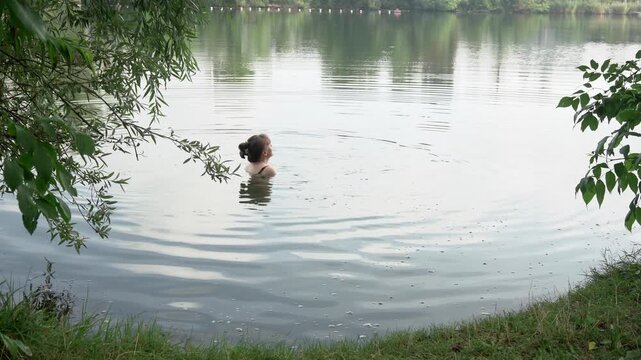 A serene medium shot of a young woman with a hair bun, sitting in a calm natural lake. She gently moves her hands through the water, creating soft ripples