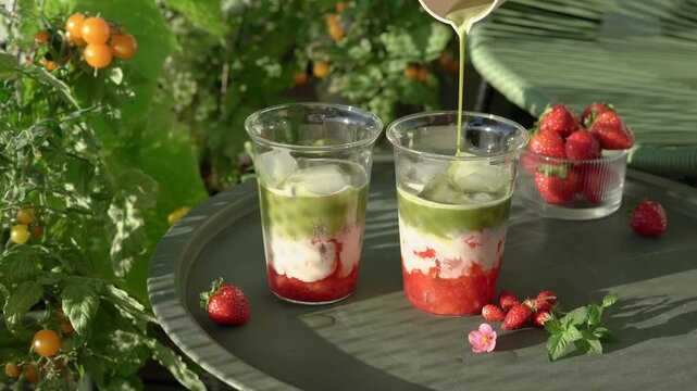 A detailed close-up shot of a refreshing layered summer drink being prepared on a terrace. Green matcha tea is gently poured from a small bowl into transparent cups filled with ice, plant-based milk