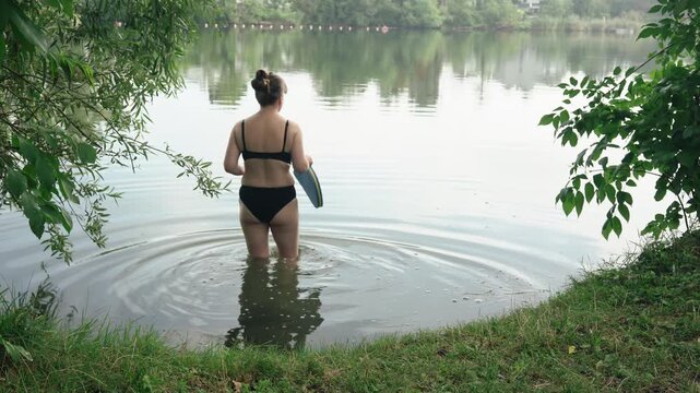 A serene medium back shot of a young woman with a hair bun, wearing a black bikini and holding swimming equipment. She slowly walks into the calm water of a natural lake on a quiet summer morning.