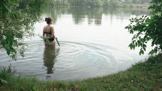 A serene back shot of a young woman with a hair bun, wearing a black bikini and holding a blue swimming kickboard. She slowly steps into the shallow water of a calm lake on a quiet summer morning
