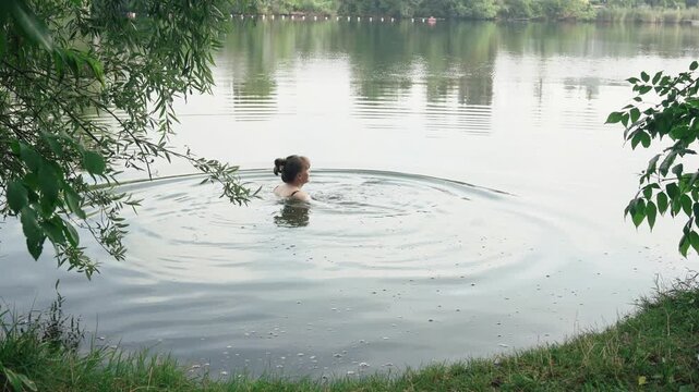 A peaceful medium back shot of a young woman with a hair bun, wearing a black bikini, sitting calmly in the shallow water of a natural lake. Beautiful soft ripples expand around her
