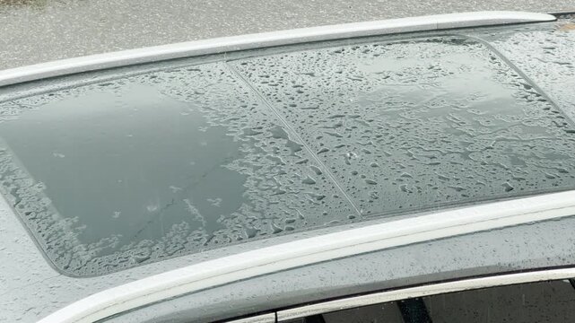 A car sunroof gathers heavy raindrops during a heavy downpour. Water droplets splash across the glass surface. This rainy scene creates a gloomy, lonely, and melancholic mood.