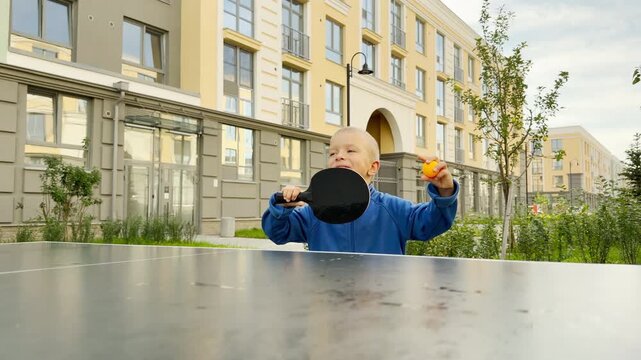A happy young boy holds a paddle and ball by an outdoor ping-pong table. He smiles cheerfully while playing in a bright residential courtyard. The joyful child feels excited.