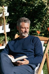 Fototapeta premium Mature man reading book in sunny garden chair for relaxation lifestyle portrait and outdoor leisure background for wellness marketing