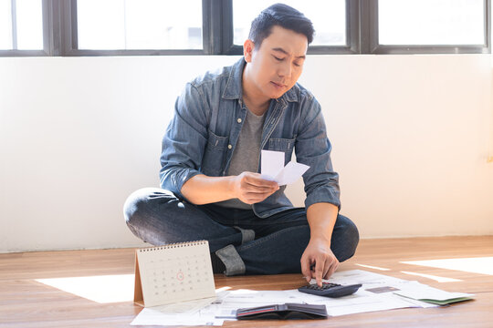 Stressed Asian man sitting on floor using calculator to check receipts and calendar for monthly expenses, financial problems, debt management, and bankruptcy concept at home.