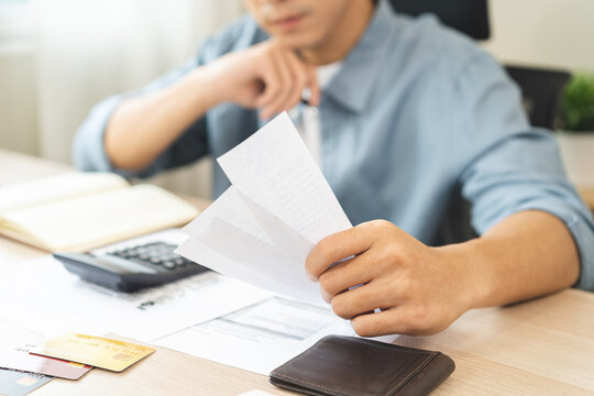 Debt and financial concept, stressed asian young man have a headache while male sitting calculate expenses, income, hand holding bills for prepare payment or pay bills on table at home.