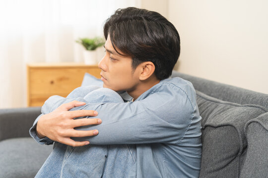 Sad unhappy, alone asian young man headache holding forehead thinking about problem, difficulty, feeling failure and exhausted, suffering sitting on sofa, grief sorrow and bad relationship or break up