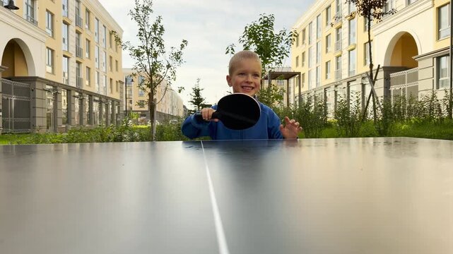 A joyful young boy plays table tennis outdoors. He smiles happily while holding a paddle near a ping pong table in a bright residential courtyard.