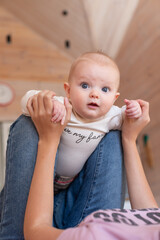 Fototapeta premium Curious blue-eyed baby sitting upright with support from a caregiver holding both hands, practicing balance and motor skills during an important early childhood development milestone at home.
