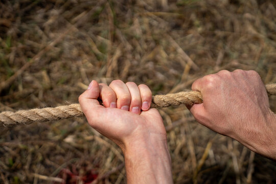 Hands of a man holding and pulling tightly a thick twisted natural jute rope outdoors, symbol of teamwork, strength, effort, and competition.