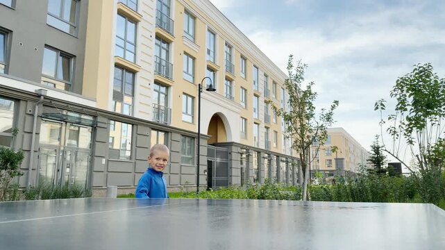 A happy young boy holds a paddle while playing table tennis. He stands in a bright courtyard near modern buildings. His gentle smile reflects pure joy and contentment.