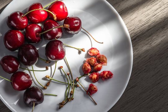  Fresh red cherries with pits and stems on a white plate in sunlight.
