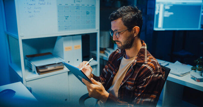 Young Caucasian male programmer use tablet for code design and plan in dark home office at night. AI technology, cybersecurity, software developer, freelance innovation, digital data network concept.