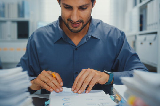 Closeup overworked young Latin businessman reading document in large stack of paperwork file at office desk. Tax audit deadline, accounting stress, corporate burnout, bookkeeping, bureaucracy concept.