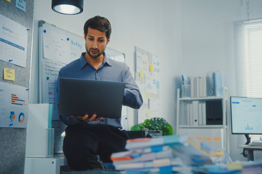 Thoughtful young Latin businessman stand with laptop analyzing data above paperwork stack. Hybrid work audit, digital transformation strategy, business corporate finance, executive decision.