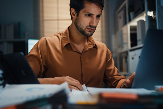 Close up overworked young Latin businessman working late on document in large stack of paper files at office night. Tax audit deadline, accounting stress, corporate burnout, urgent business concept.