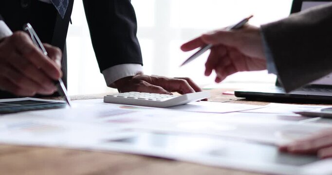 Business team analyzing financial reports and charts at a wooden table, pointing at a laptop screen, calculating figures on a calculator and planning growth strategy together