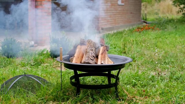 Wood burns with bright flames and rising smoke inside a metal fire pit on green grass near a country house, preparing an outdoor fire for barbecue cooking. Close up.