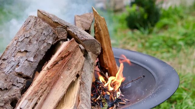 Wood burns with bright flames and rising smoke inside a metal fire pit on green grass near a country house, preparing an outdoor fire for barbecue cooking. Close up.
