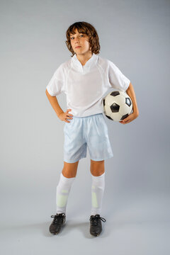 Young male soccer player standing in his uniform with a soccer ball under one arm and his hand on his hip