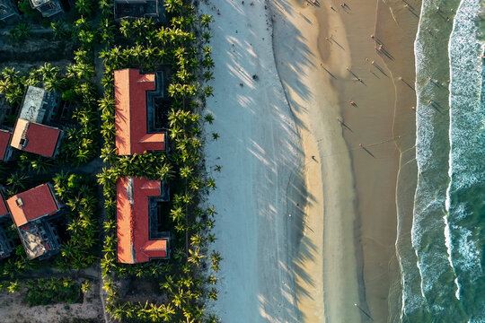 People walk along the sandy shoreline near tropical resort buildings and lush green palm trees at Cua Dai Beach in Hoi An during the bright afternoon
