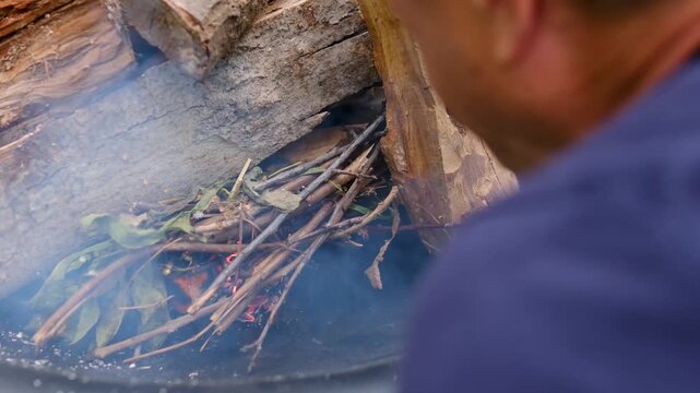 carefully blows on burning paper beneath thin dry twigs to strengthen the flame and start a wood fire in an outdoor fire pit for barbecue preparation. Close up.