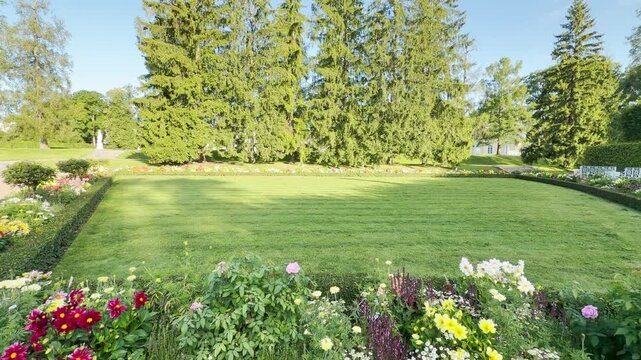 Bright, colorful flowers bloom around a neat green lawn in Catherine Park, Saint Petersburg. Tall evergreen trees stand under a clear sky, inspiring peaceful joy and serene happiness.
