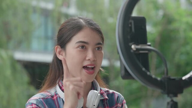 Young woman talks to a smartphone camera outdoors while wearing headphones, using expressive gestures during a natural on-camera presentation for digital media and social content.