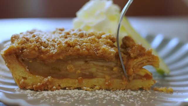 Close up of fork cutting into a delicious slice of apple crumble pie dusted with powdered sugar. Homemade sweet dessert with whipped cream. Shot with selective focus. Bakery pastry concept.