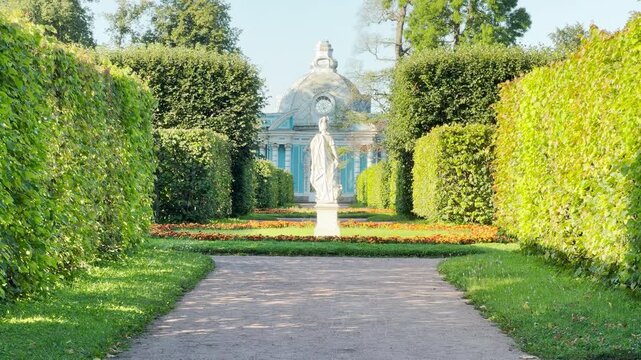 A marble statue stands gracefully along a path lined with tall hedges. A blue dome rises peacefully behind it. This scene in Catherine Park, Saint Petersburg brings wonder and serene joy.
