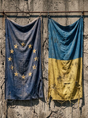 Naklejka premium tattered torn flags of european union and ukraine hang side by side on a metal rod against a cracked concrete wall reflecting a difficult period crisis and consequences of prolonged conflict