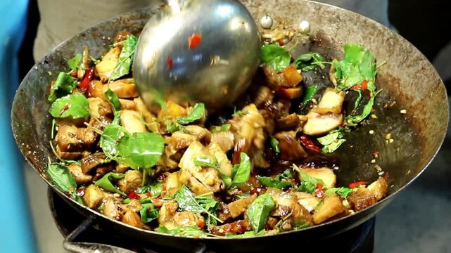 A wok sizzles with golden caramelized stir-fried pork chunks mixed with fresh aromatic holy basil and bright red chili peppers at a Thai street food night market stall in Thailand.