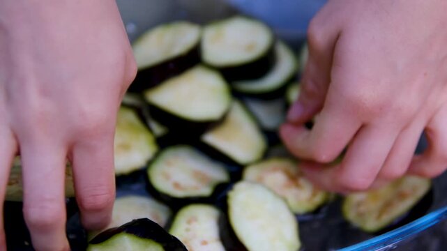 Female hands prepare eggplants for outdoor grilling by slicing, salting, rinsing and squeezing out excess moisture in a glass bowl before cooking on barbecue. Close up.