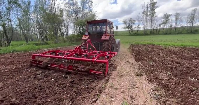 Red tractor pulling harrow in spring field prepares soil for planting on cloudy day.
