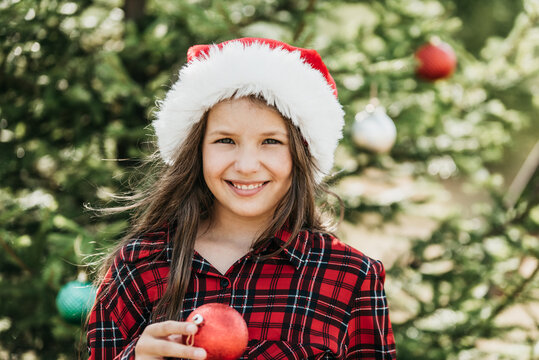 Summer Christmas with girl wearing Santa hat holding red Christmas ornaments beside decorated evergreen tree in sunny summer garden.