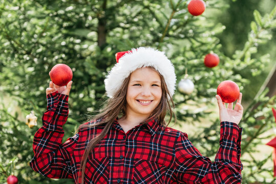 Summer Christmas with girl wearing Santa hat holding red Christmas ornaments beside decorated evergreen tree in sunny summer garden.