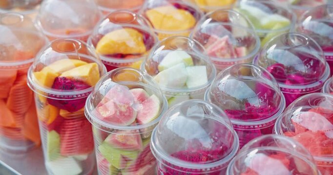 Sliced tropical fruits including dragon fruit, mango, and guava in plastic cups displayed at an outdoor street market, healthy snack retail, food stall background.